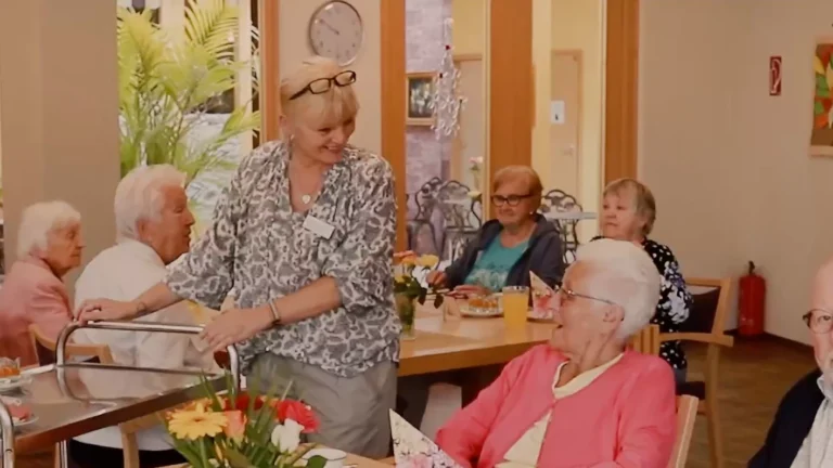 Das Bild zeigt eine herzliche Mitarbeiterin der Betreuungsstuben, die einer Seniorin auf einem schön gedeckten Tisch mit Blumen eine Mahlzeit serviert.