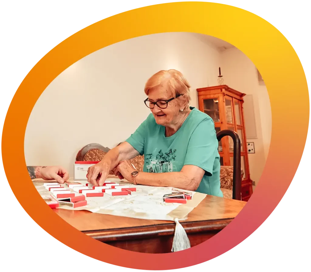 Klientin mit einem Mitarbeiter des Betreuungsdienstes bei einem Memory-Match in einem gemütlich eingerichtetem Wohnzimmer.